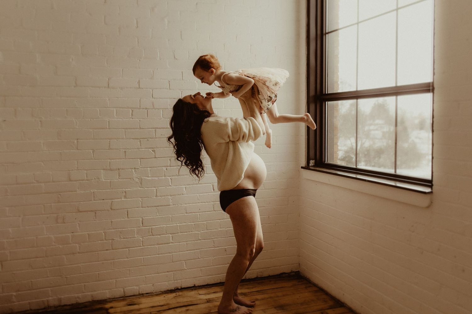 Pregnant mom taken in a studio near a large window while she catches her little girl in her arms by Bowmanville family photographer
