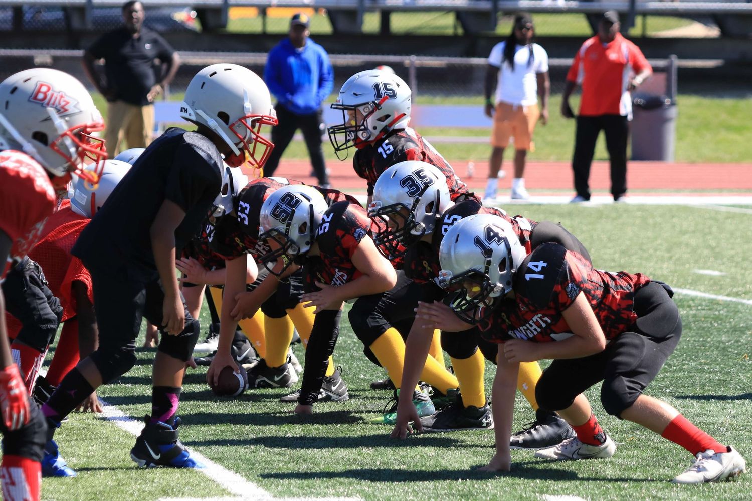 Youth football teams line up at scrimmage during a game on an artificial turf field.