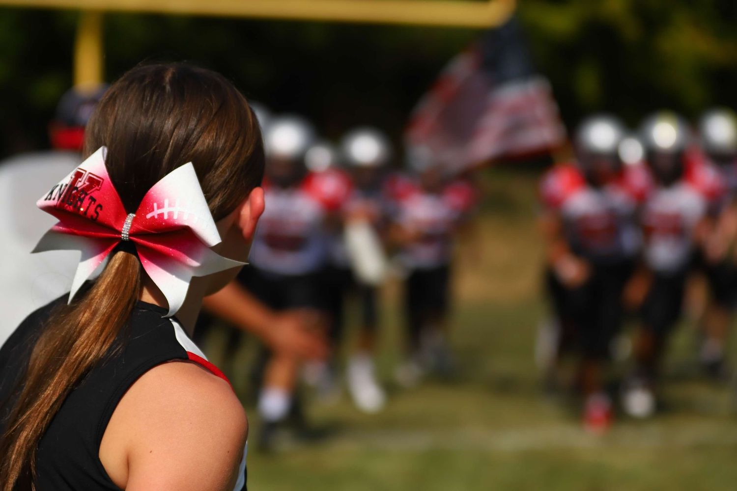 Group of cheerleaders perform on a sunny day at a sporting event with matching red bows and uniforms.