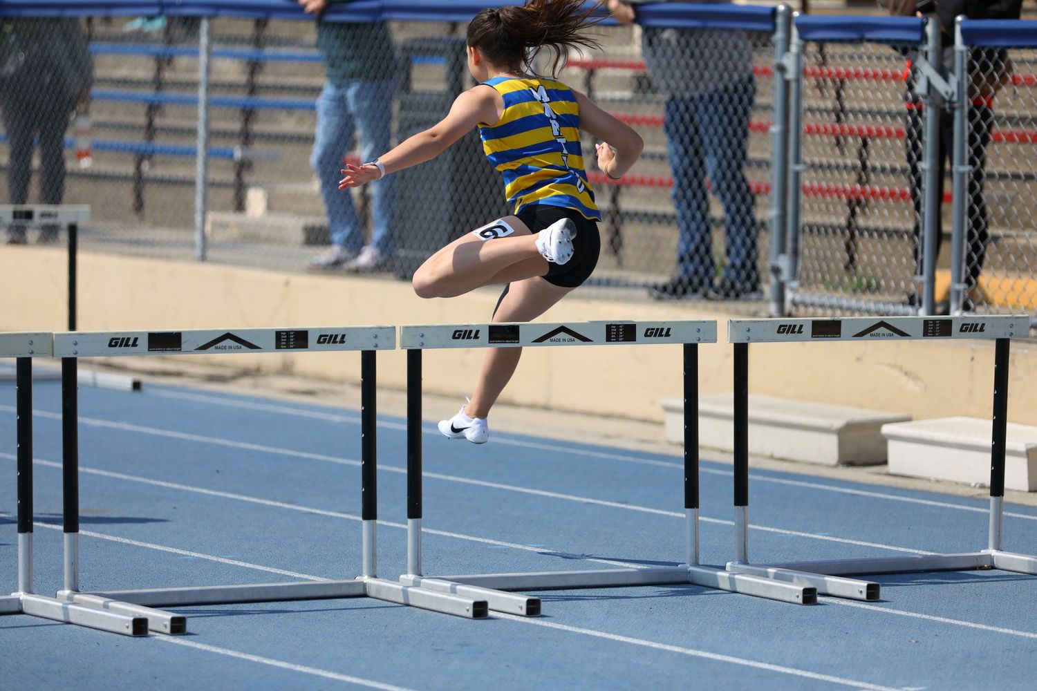 Athletes competing in a track and field hurdles event on a blue running track at an outdoor stadium.