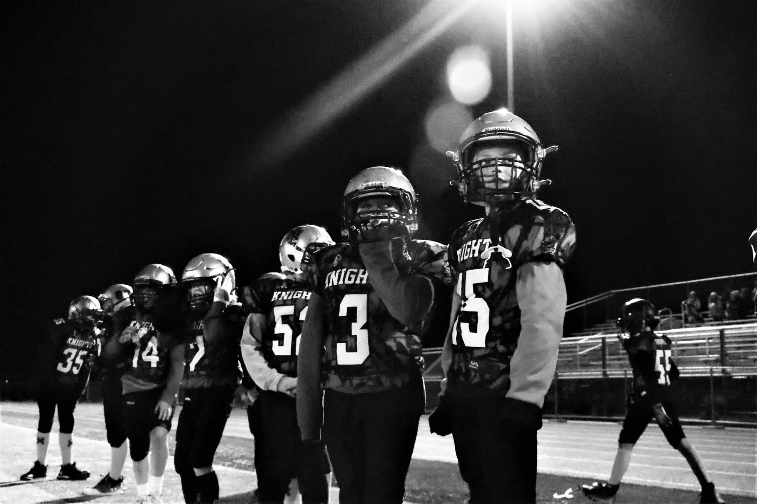 Football players facing off in a dramatic black and white shot under stadium lights at night.