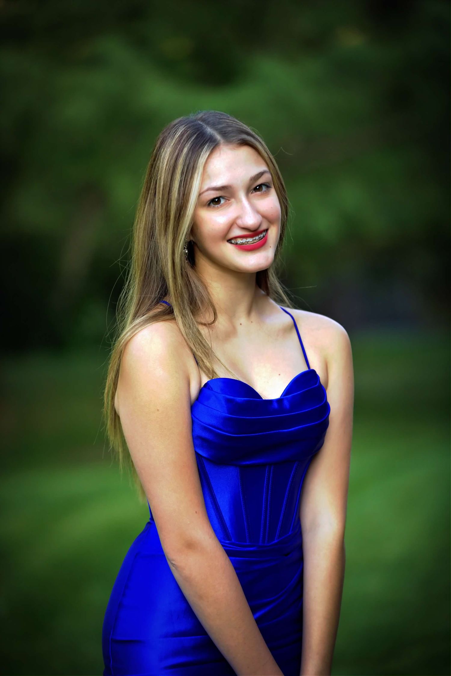 Senior portrait series featuring woman in royal blue velvet dress against natural bokeh background.