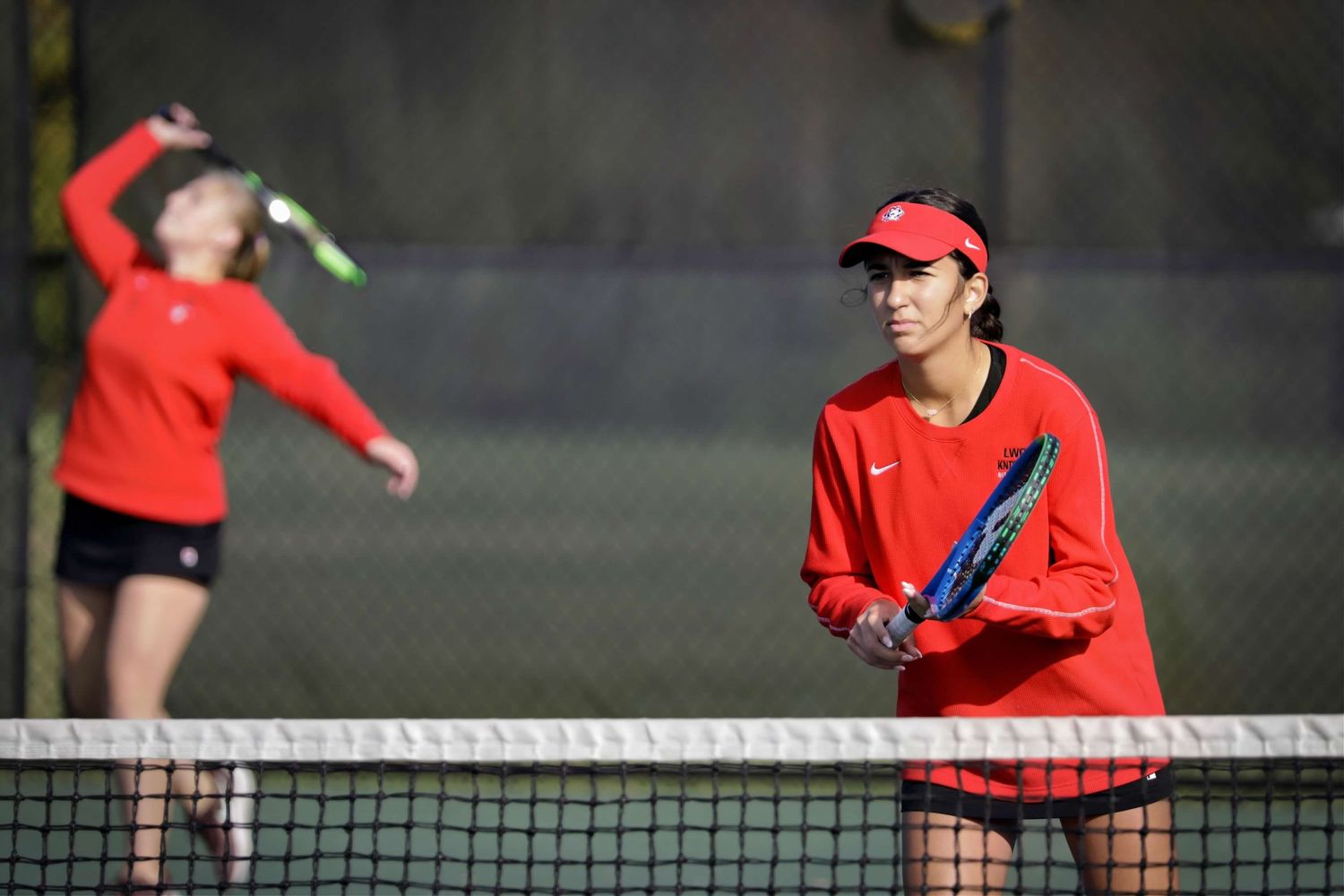 Tennis player in bright red shirt serves during competitive outdoor match on hard court surface.