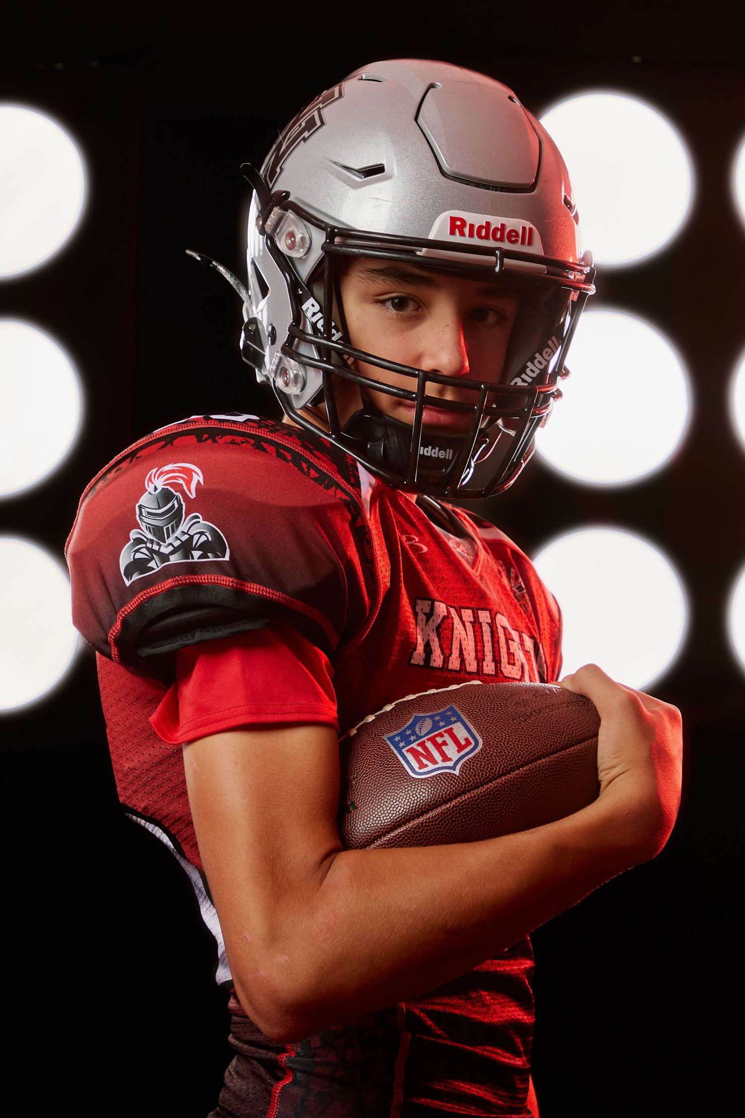 Football player in red jersey and helmet posing with football against dark background with dramatic lighting effects.