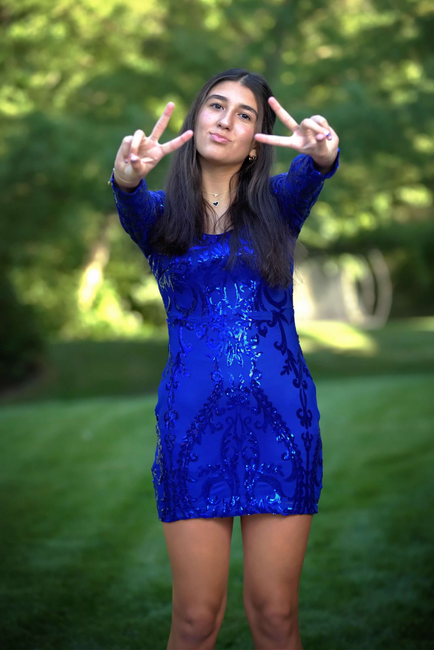 Person wearing vibrant royal blue sequined outfit poses playfully in outdoor garden setting with peace signs.