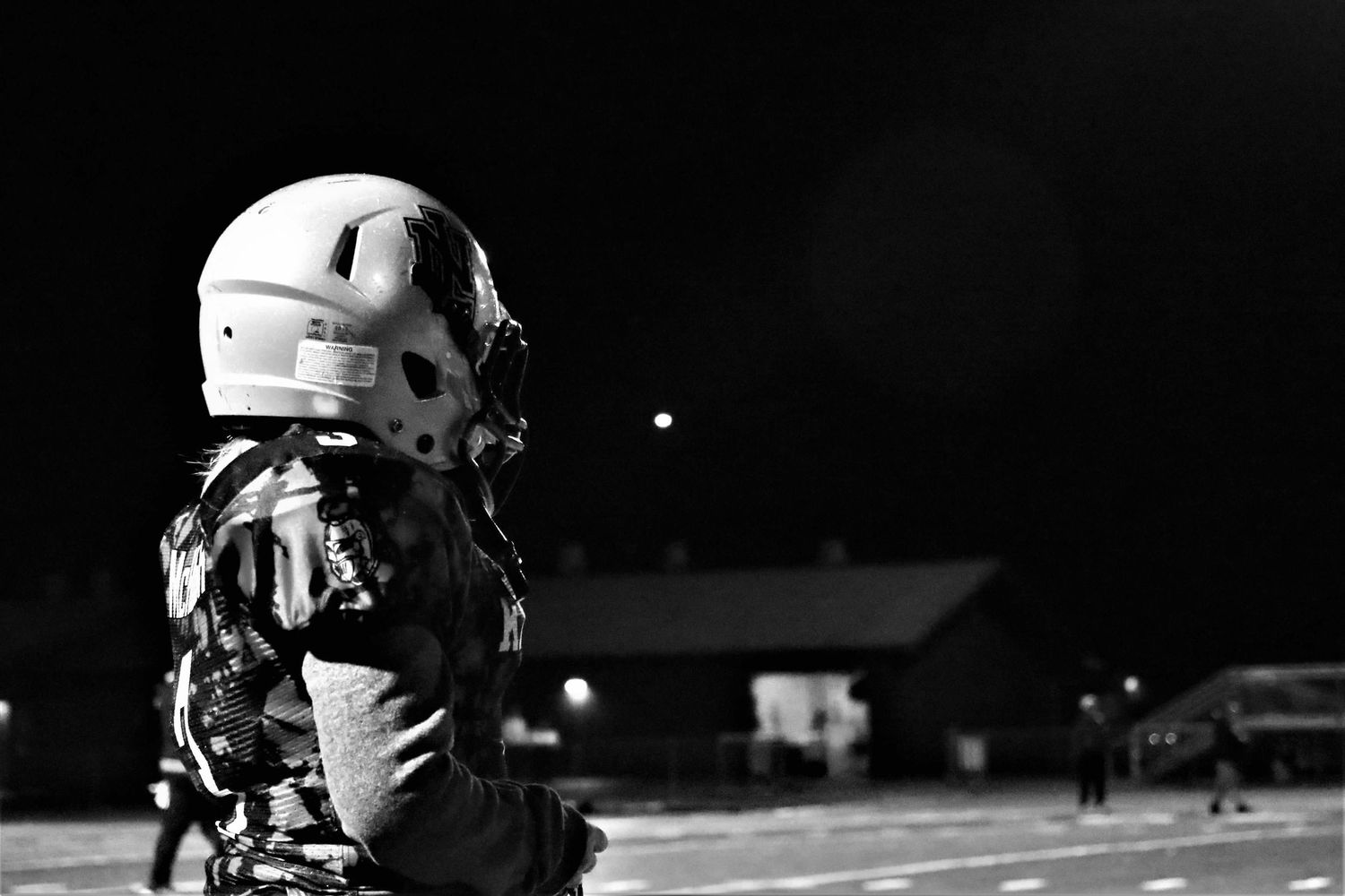 Night football action shot shows players in white and dark uniforms lined up at the line of scrimmage.