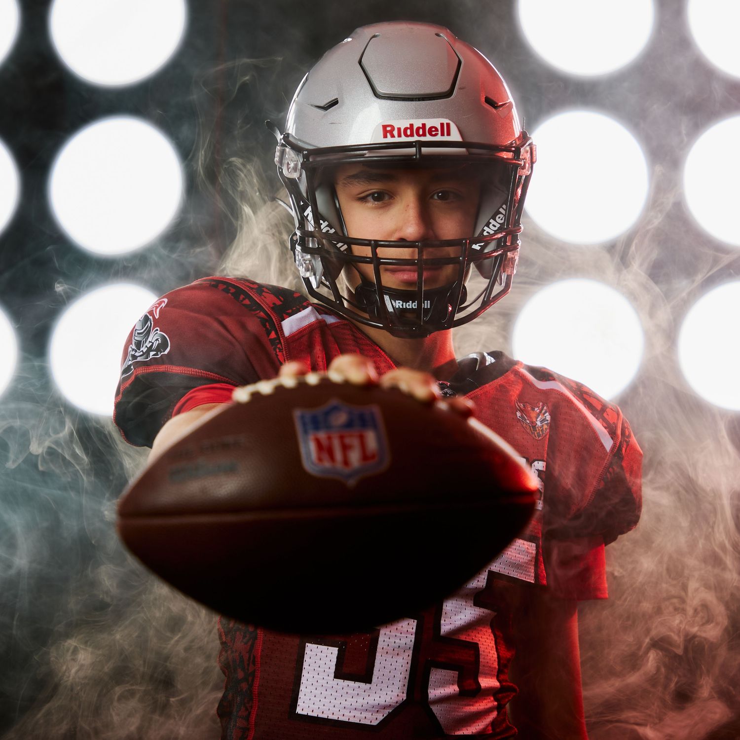 Football player in red uniform and grey helmet holding ball against dramatic spotlit background.