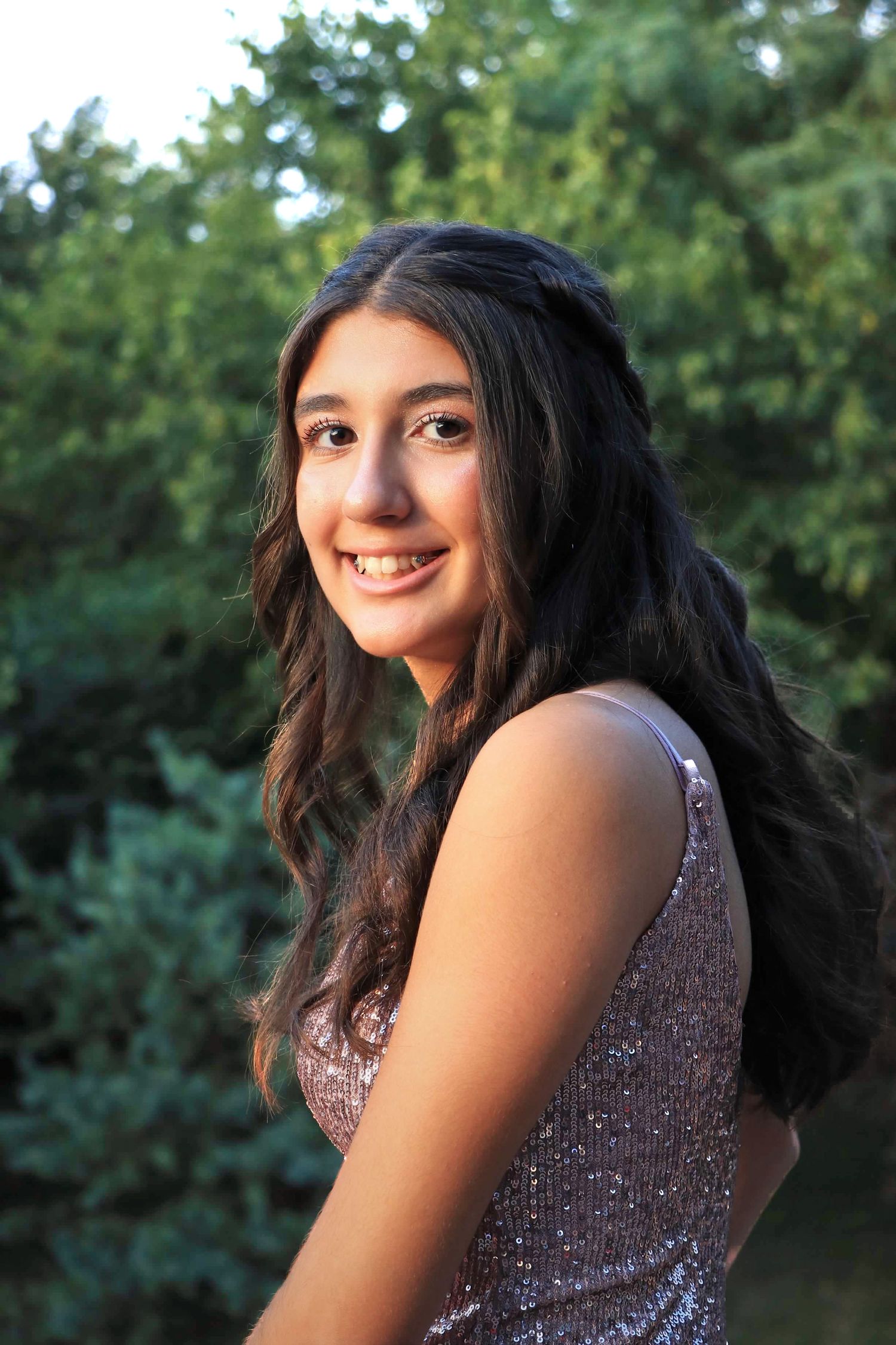 Person wearing a sparkly purple top smiling against a green outdoor background filled with trees and foliage.