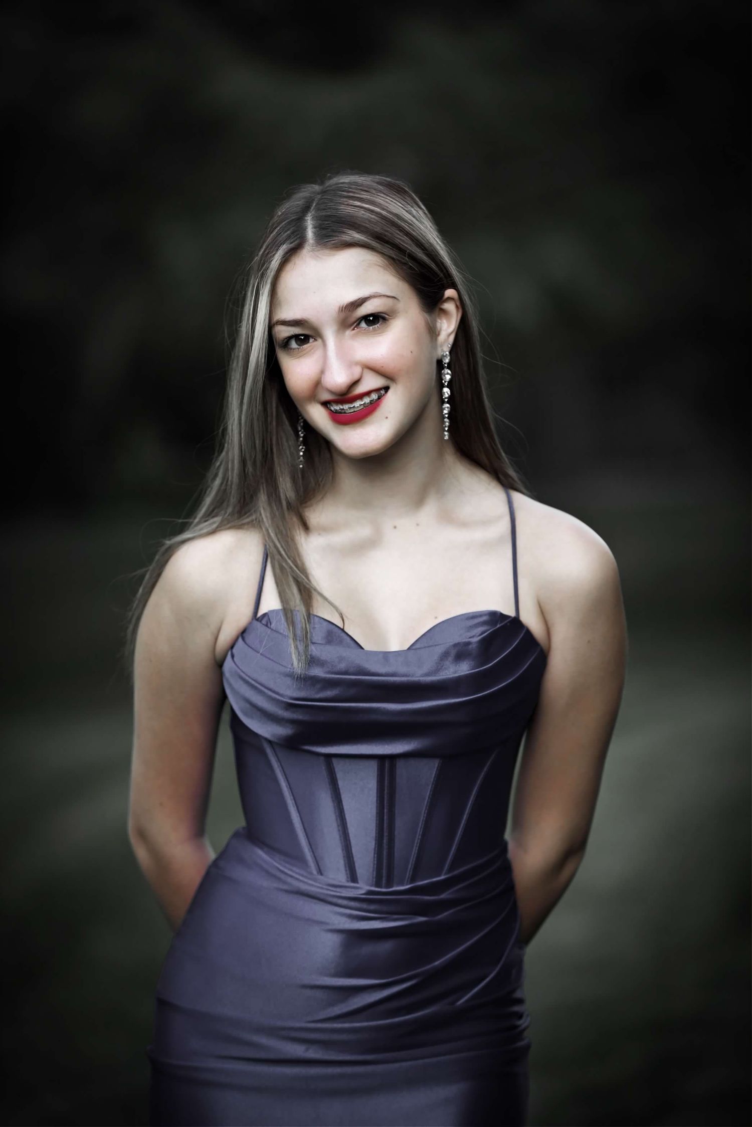 Person in dark satin evening dress poses elegantly against blurred dark background.