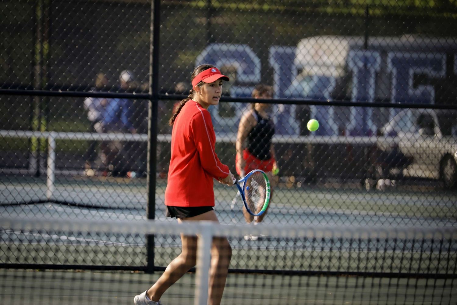 Players compete in a tennis match on outdoor courts during daytime with spectators watching from the sidelines.