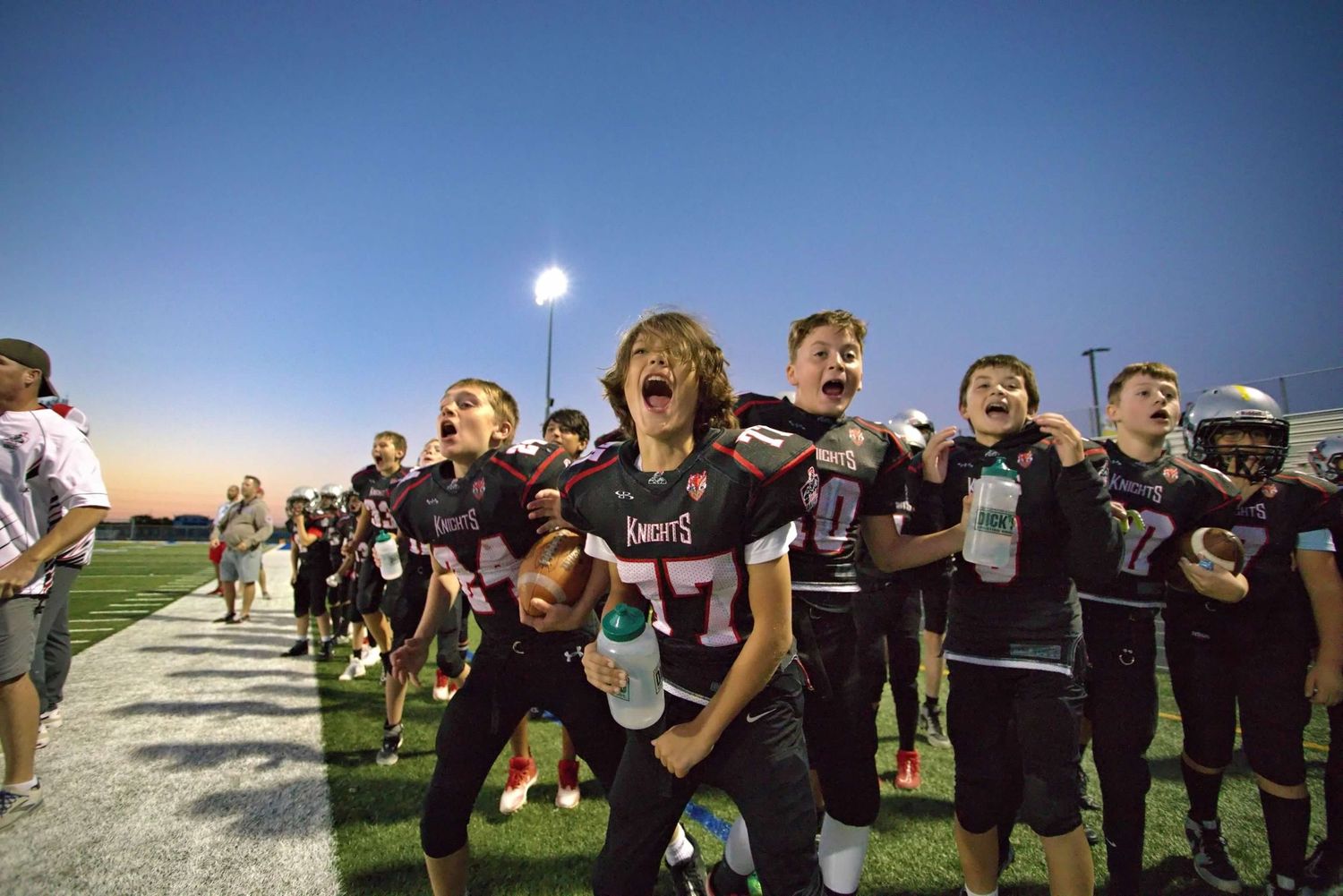 High school football team celebrating on field with arms raised in victory at sunset.