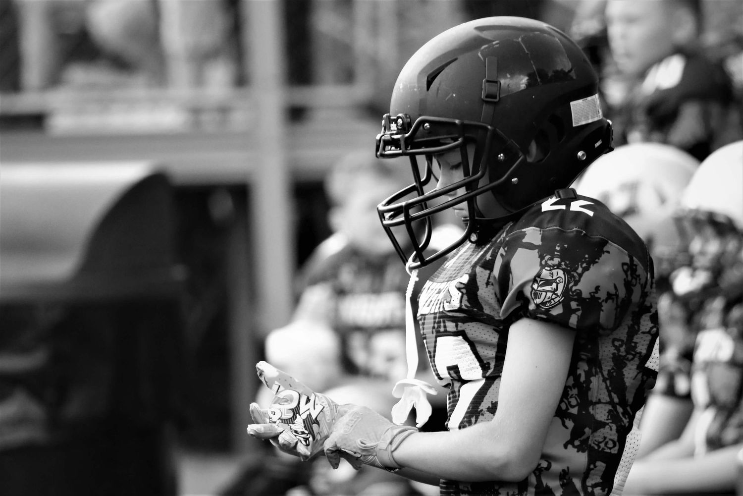 Black and white image showing a row of football players in helmets on the sideline during a game.