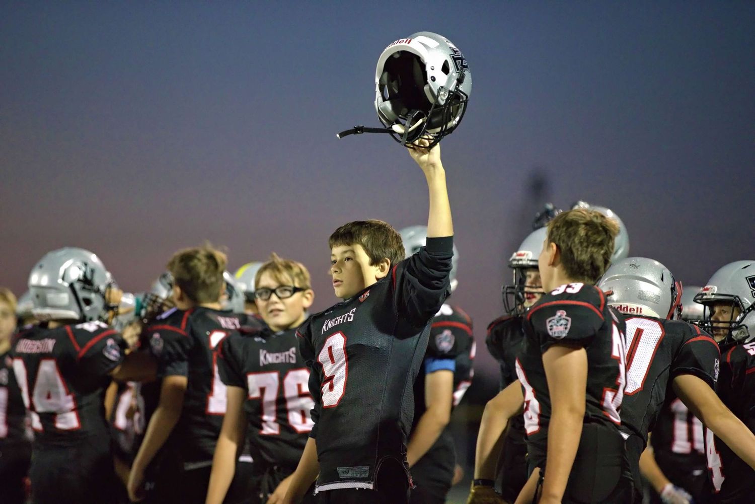 Football players raise their helmets in unison against dark blue sky during night game.