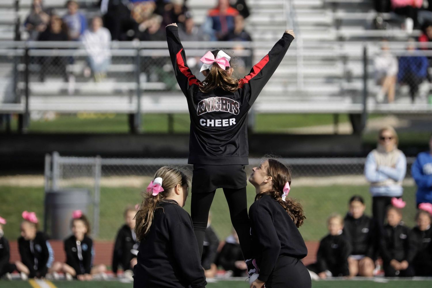 Cheerleaders perform synchronized high kicks during an outdoor sporting event on a field with spectators in the background.