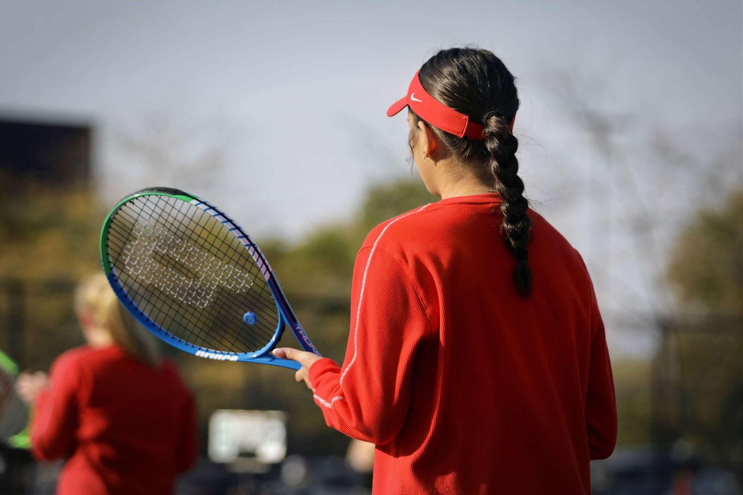 A tennis player in red attire with a braided hairstyle holds a blue racket during practice.