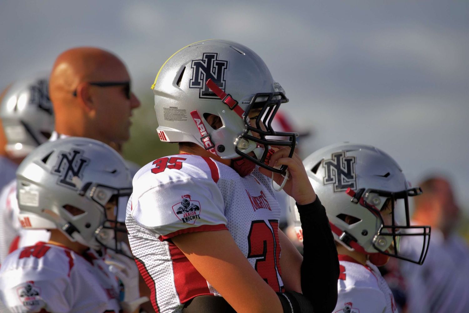 Row of football players wearing white helmets with red trim standing on sideline during game.