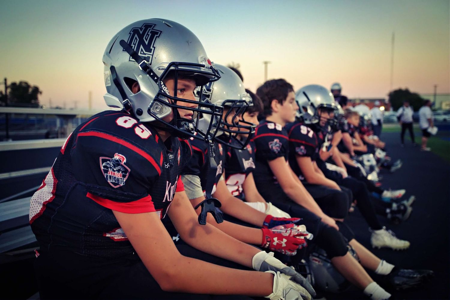 Football players in helmets and uniforms lined up on sideline at dusk during game.