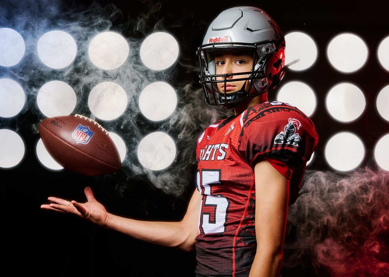 Football player wearing red jersey and helmet catches a football during a dramatic nighttime game with stadium lights.