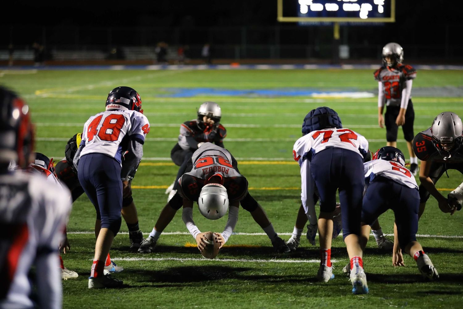 Football players engage in a nighttime game on an illuminated field with players lined up in formation.