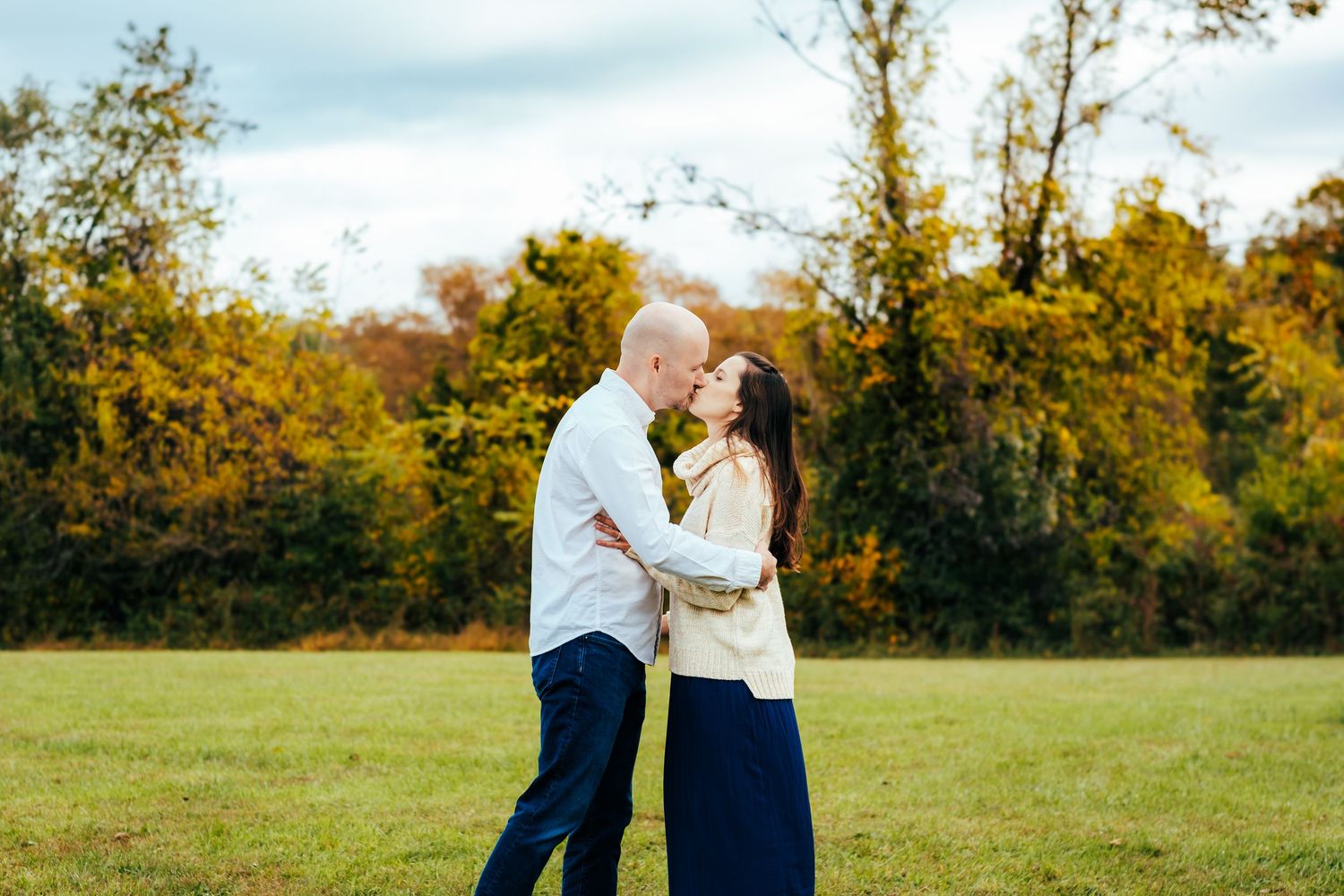 Couple sharing a romantic kiss in an open green field surrounded by colorful autumn trees during fall engagement photo session.
