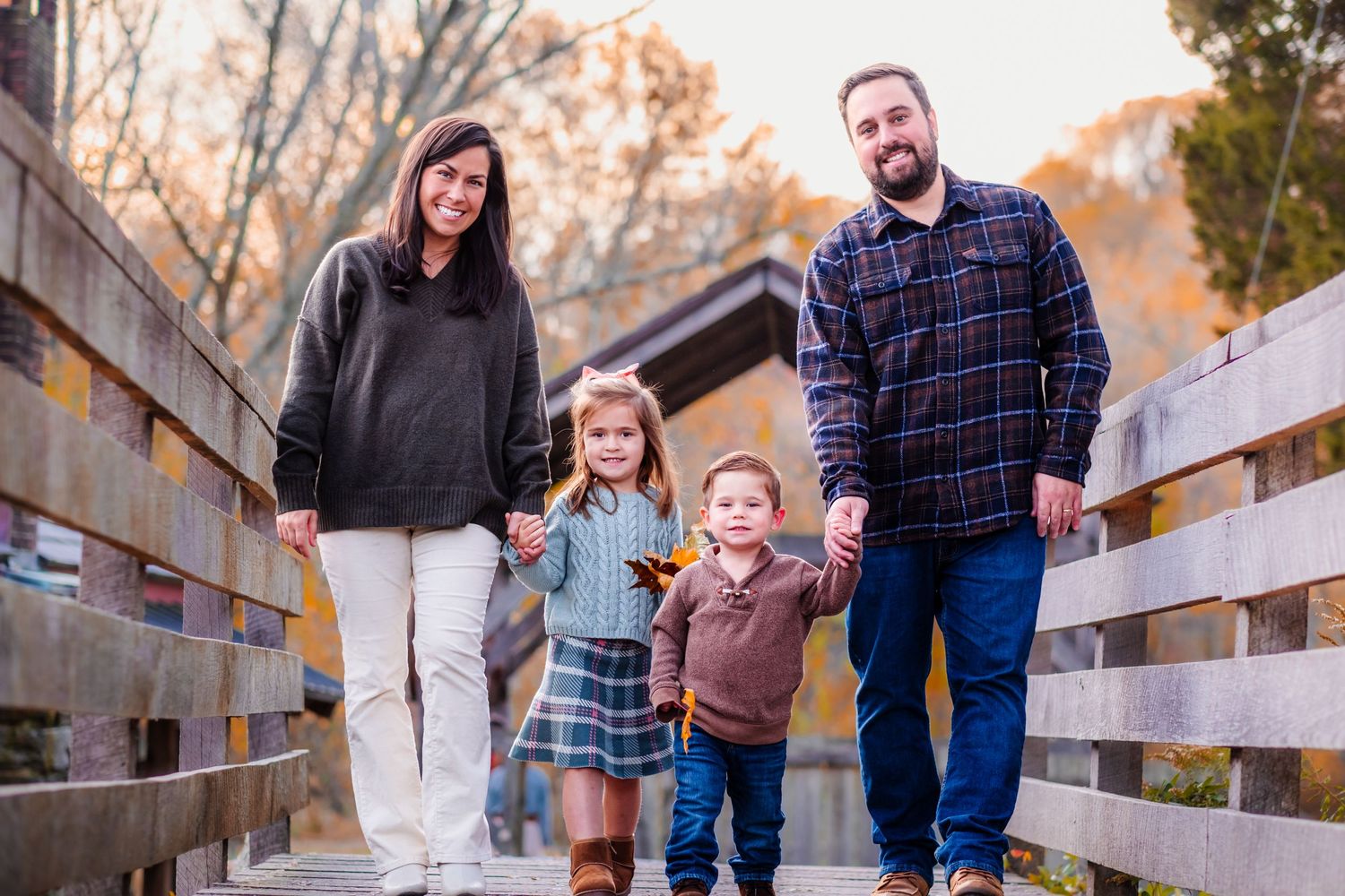 Family of four holding hands on a wooden bridge surrounded by autumn foliage for a fall family portrait.