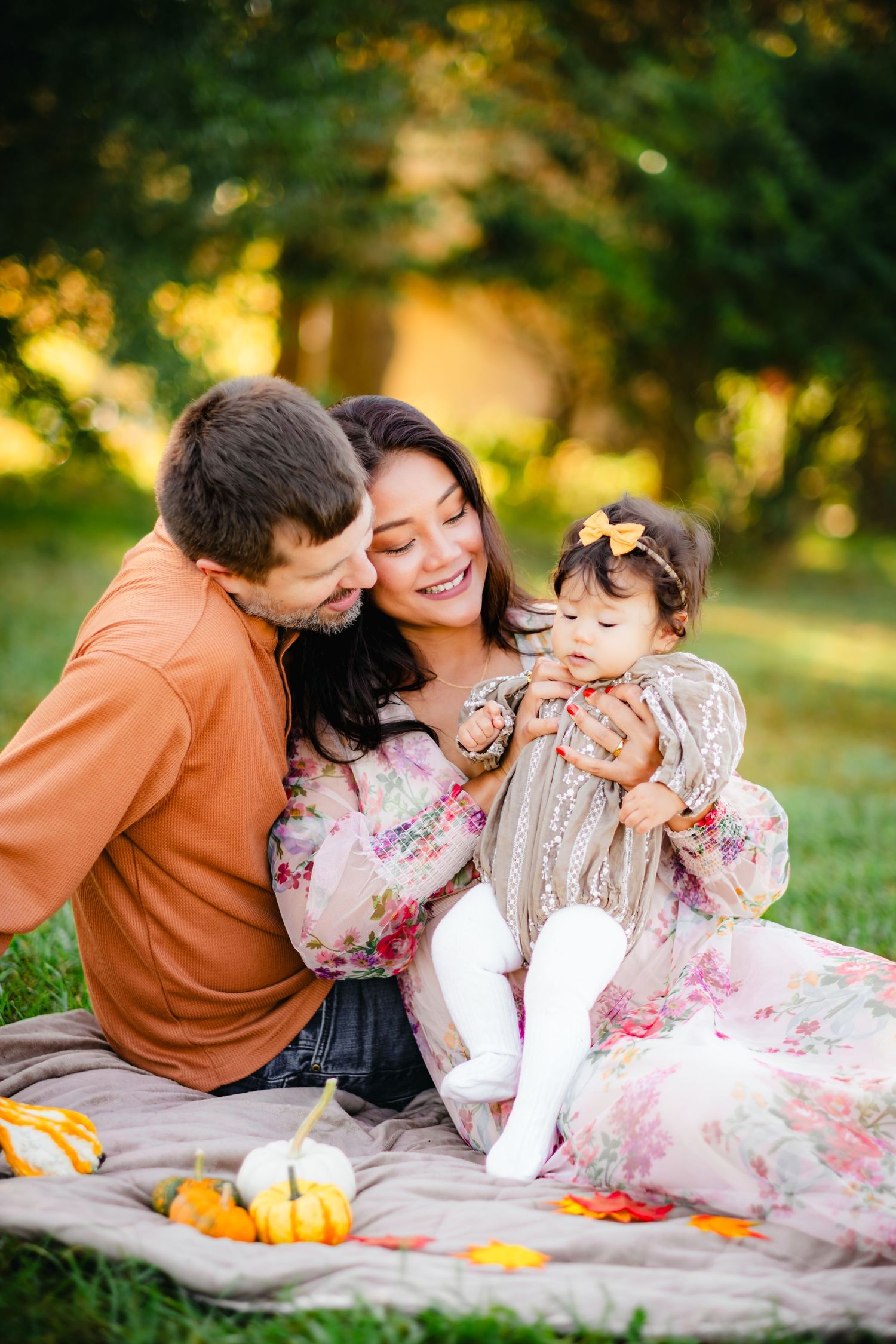 Family with baby girl sitting on blanket in autumn park with pumpkins during golden hour outdoor photo session.