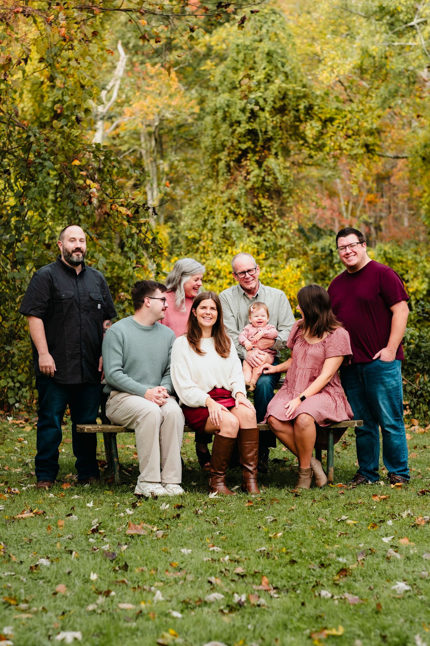 Family of eight poses outdoors on a bench surrounded by colorful autumn foliage for a fall portrait session.