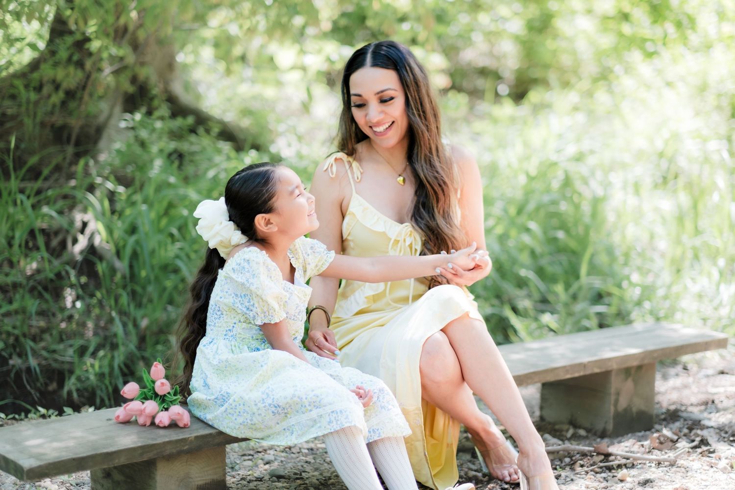 Mother and daughter in pastel dresses sitting together on a bench in a garden.