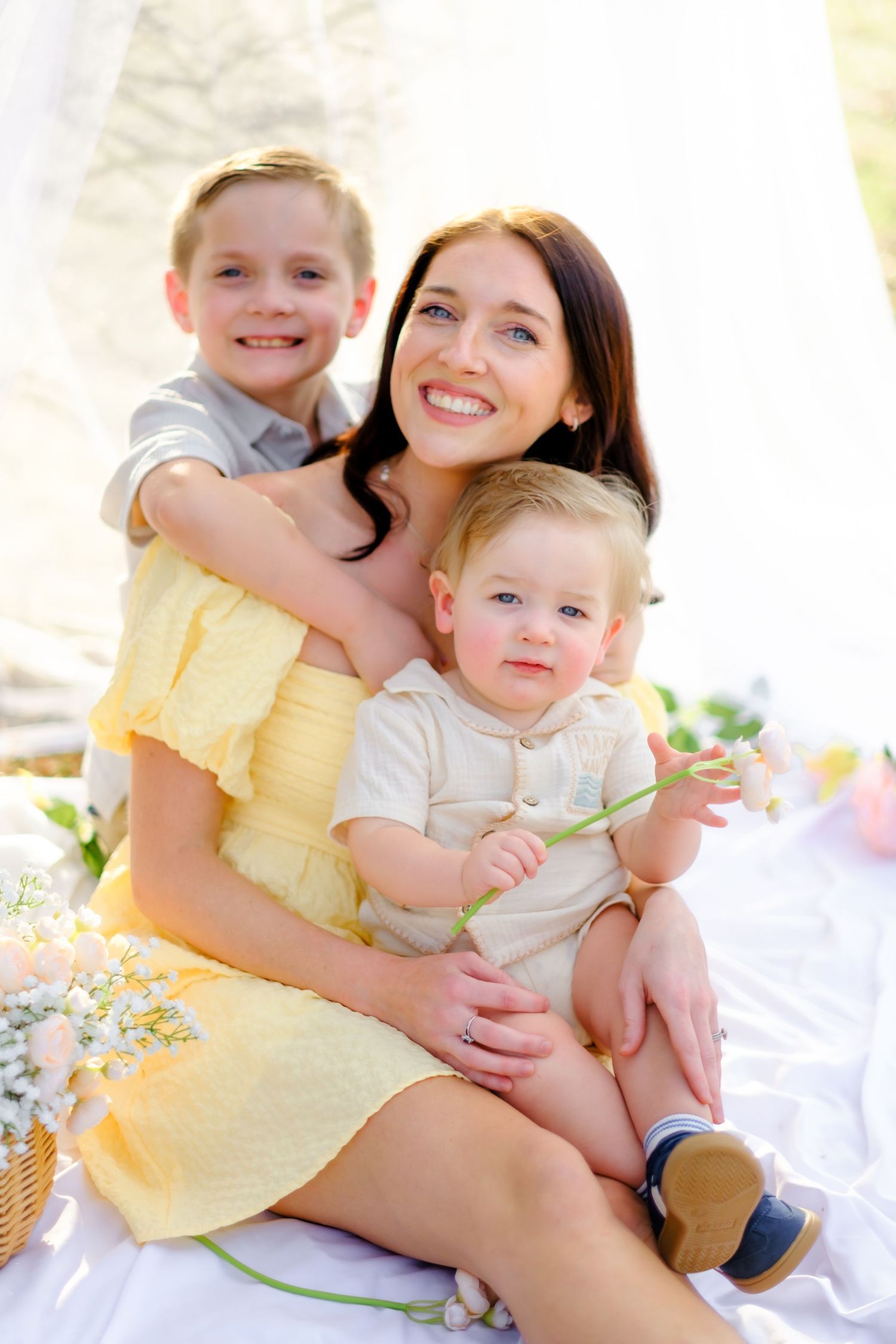Smiling mother in yellow dress poses with two young sons on white blanket during sunny outdoor photoshoot.