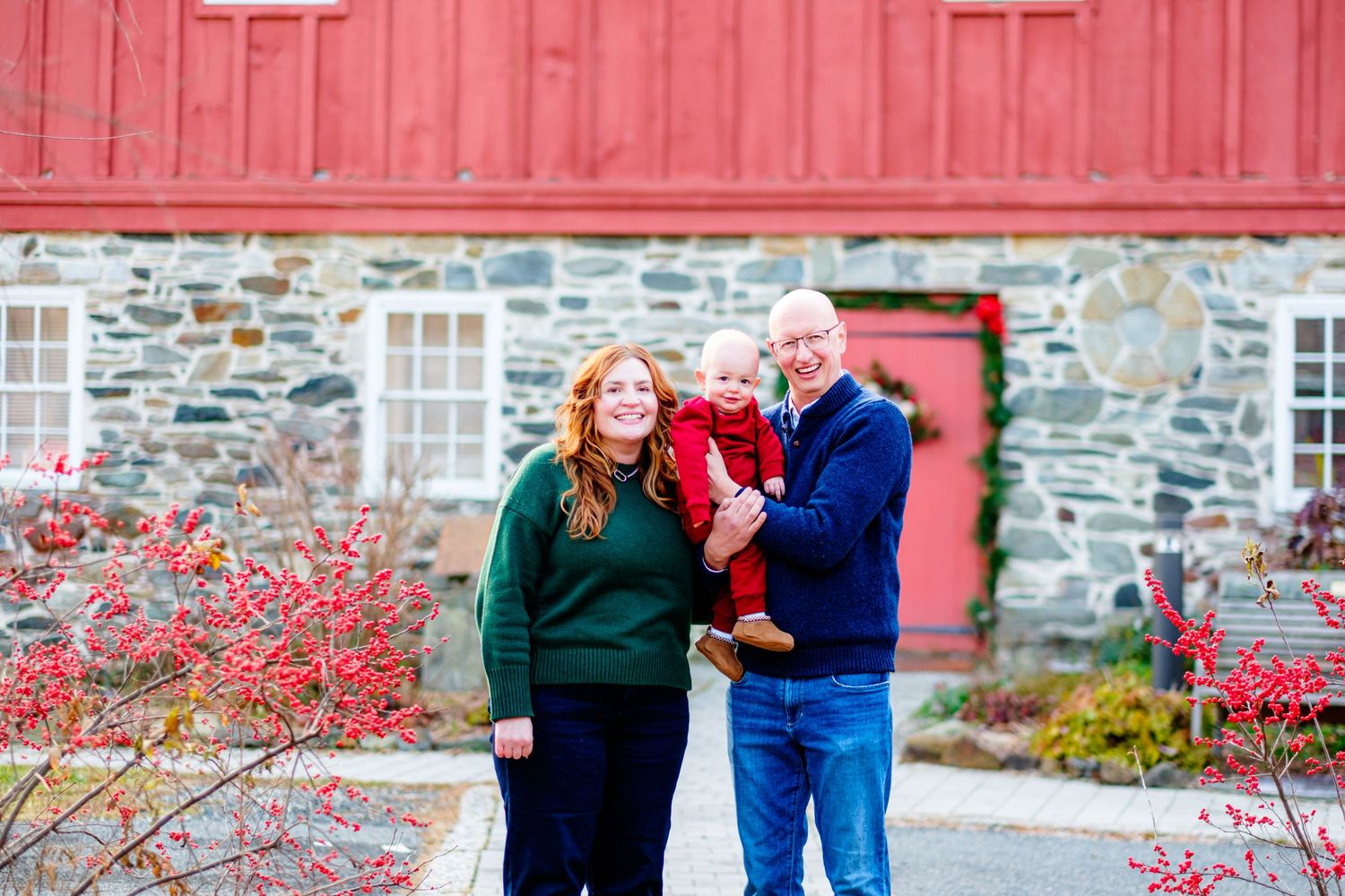 Family of three posing outdoors near a rustic stone building with red barn doors and vibrant red bushes in autumn.