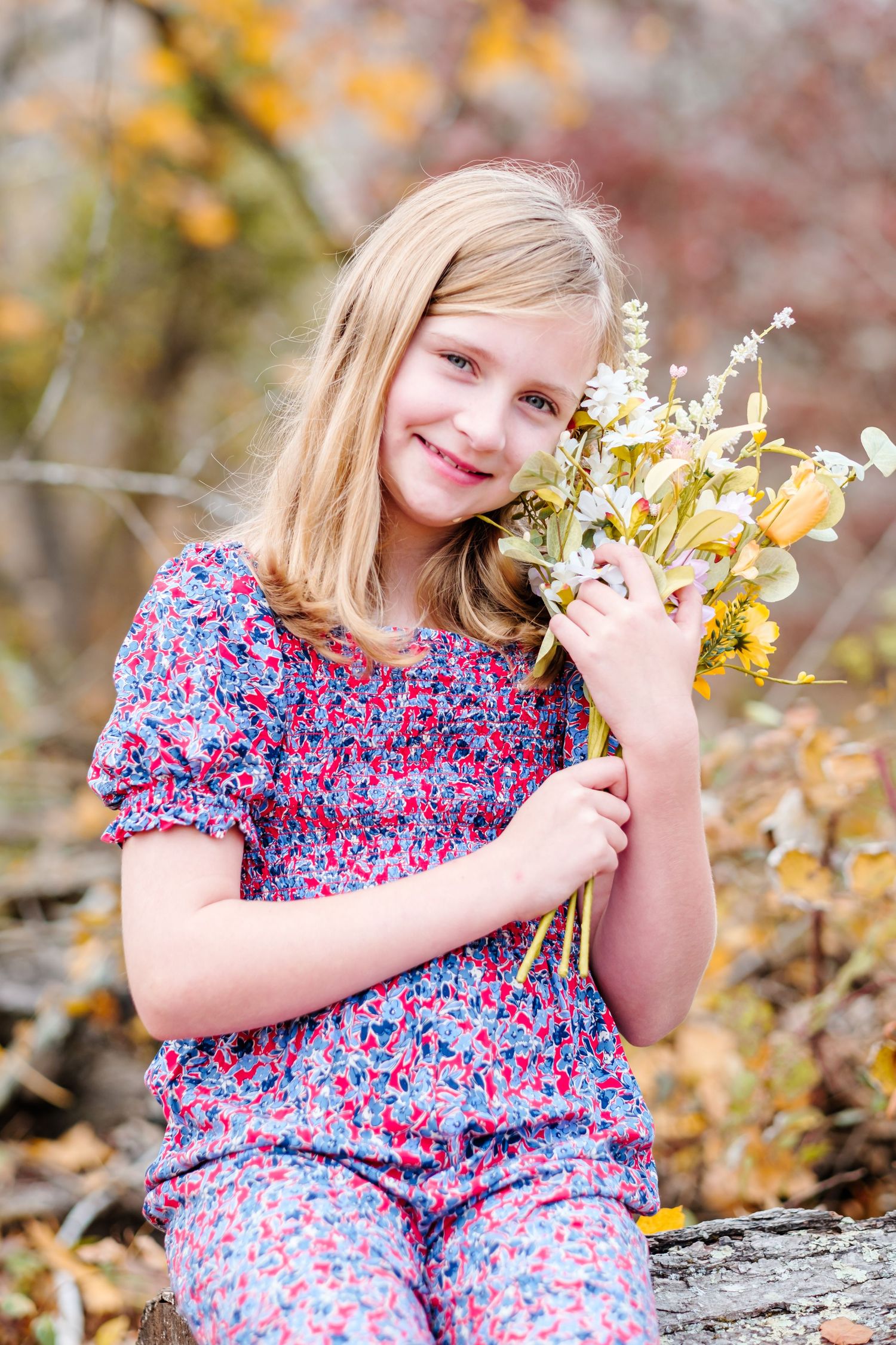 Smiling blonde girl in floral outfit holding wildflowers outdoors in autumn setting.
