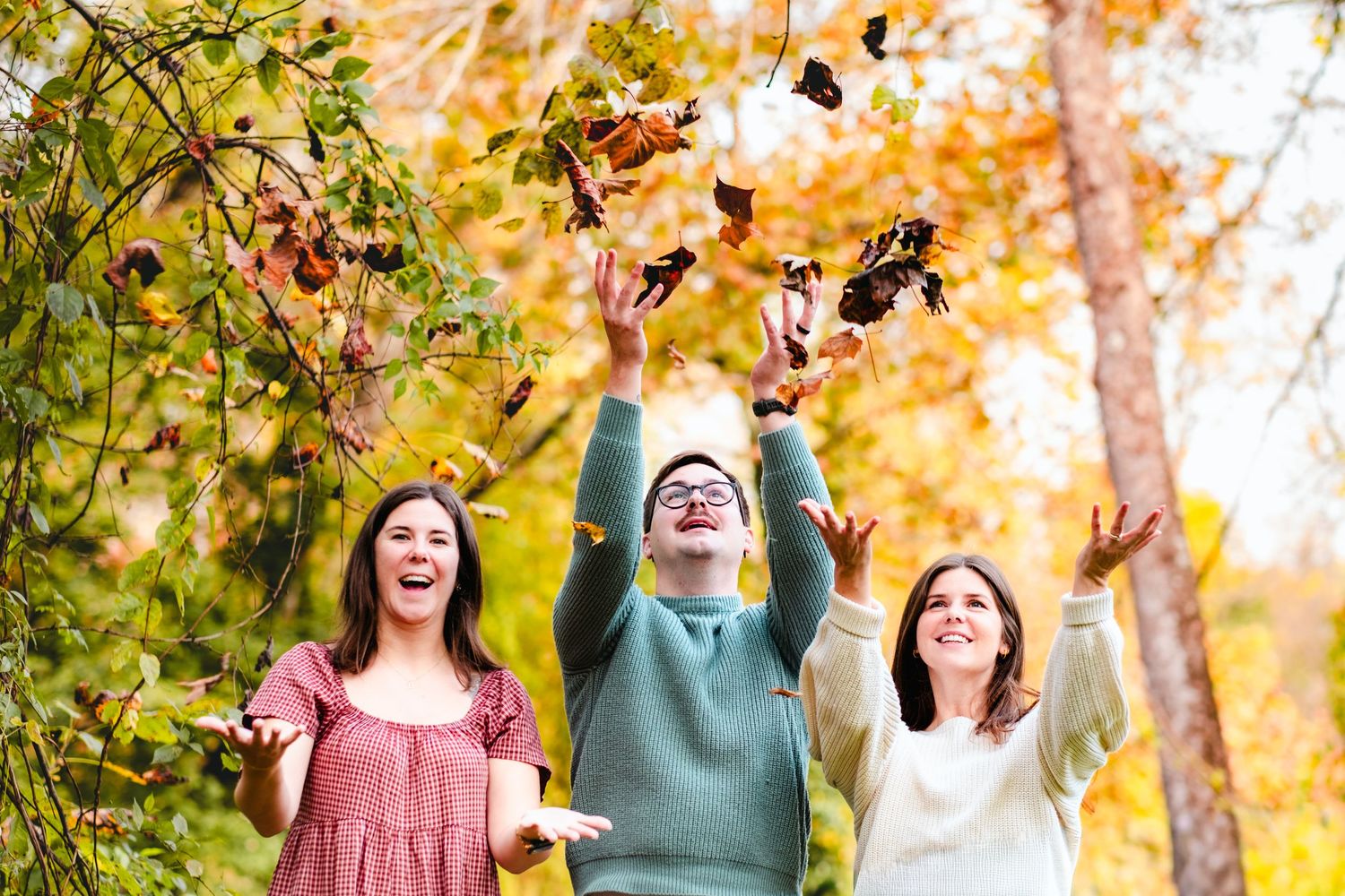 Three joyful young adults tossing autumn leaves in a colorful fall forest setting.