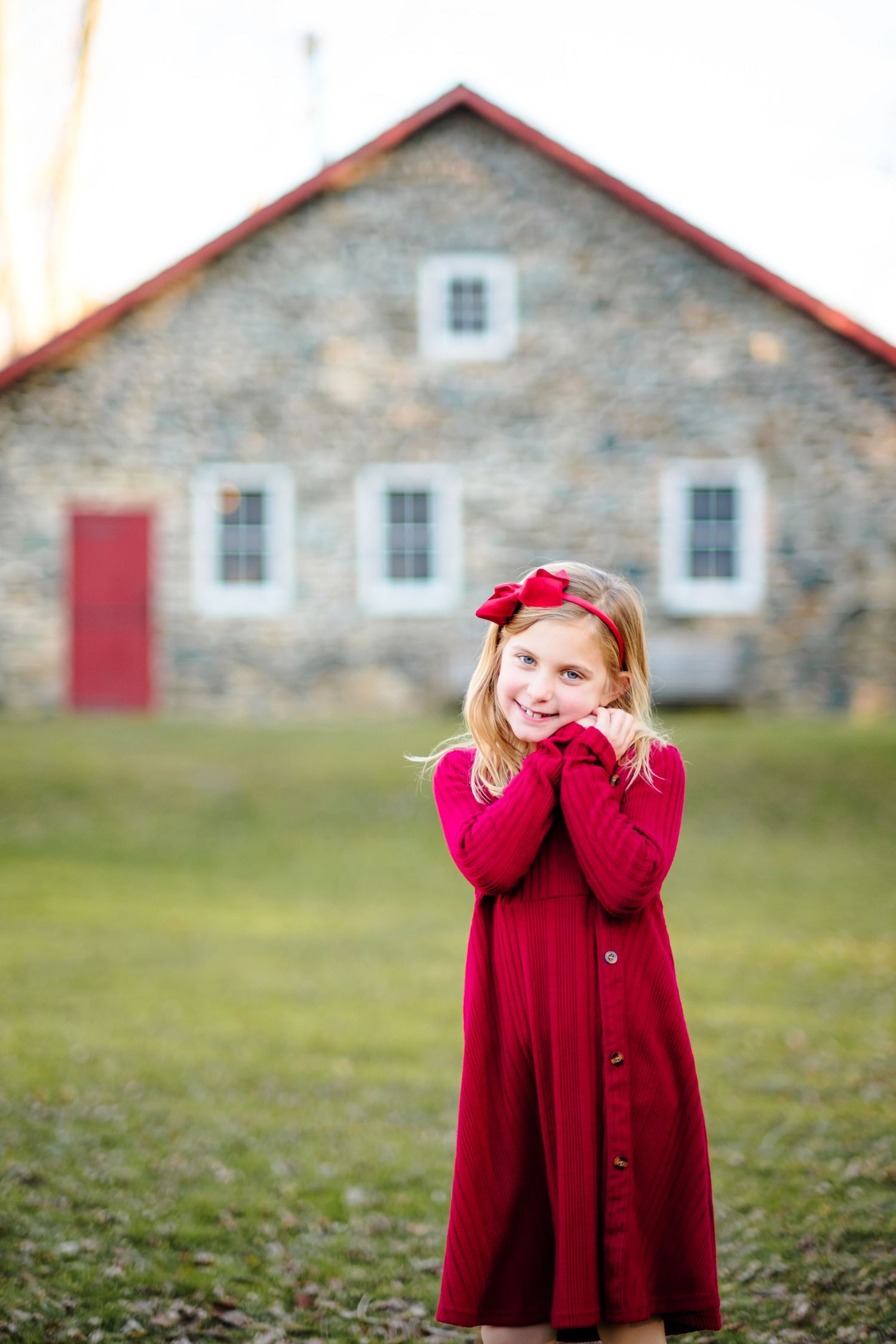 Young girl in red dress and bow smiles outdoors near rustic stone building on green lawn.