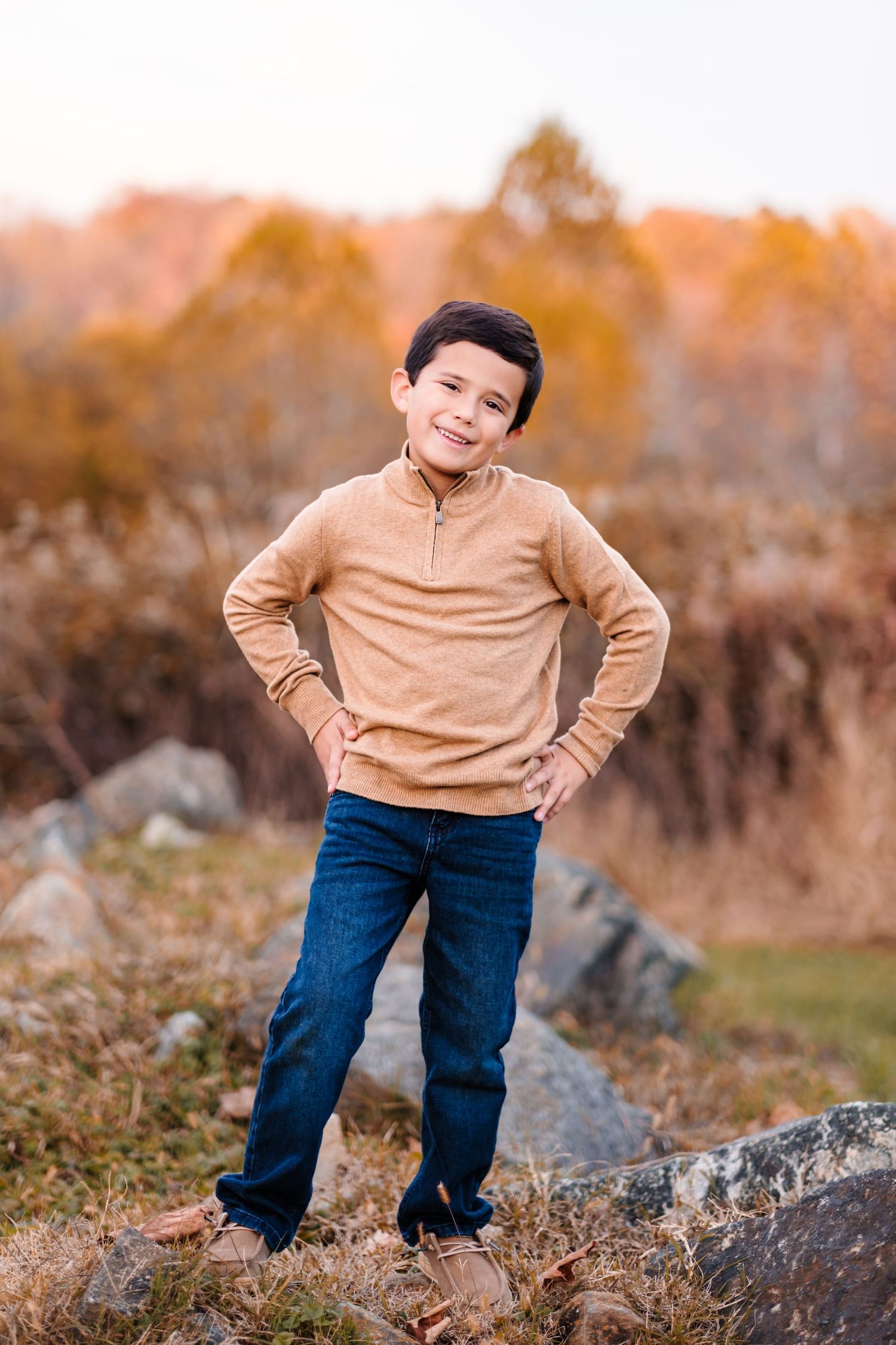 A young boy in a tan sweater and jeans poses with hands on hips outdoors in an autumn setting.