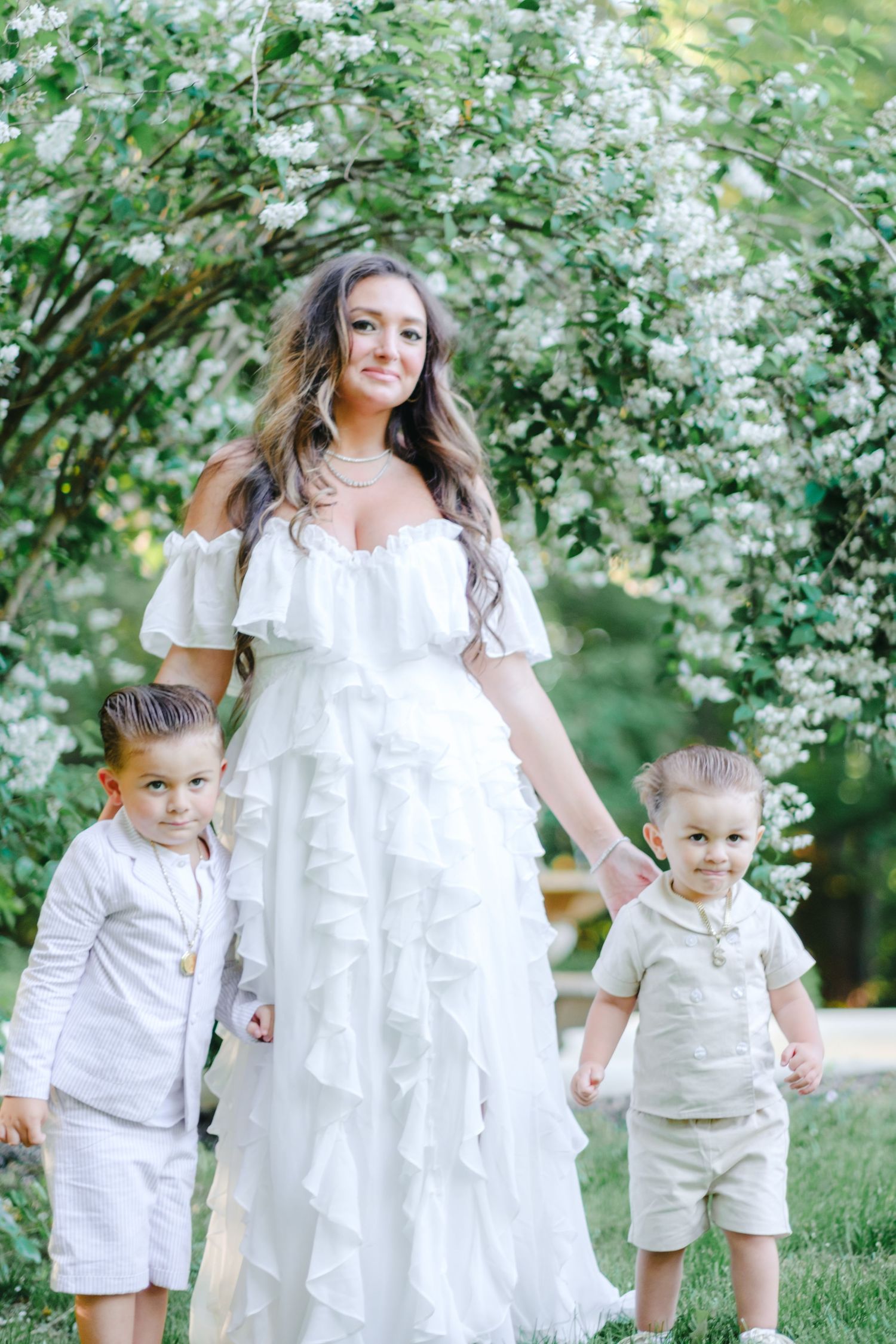 A woman in a white ruffled dress holds hands with two young boys in light outfits under a flowering arch outdoors.