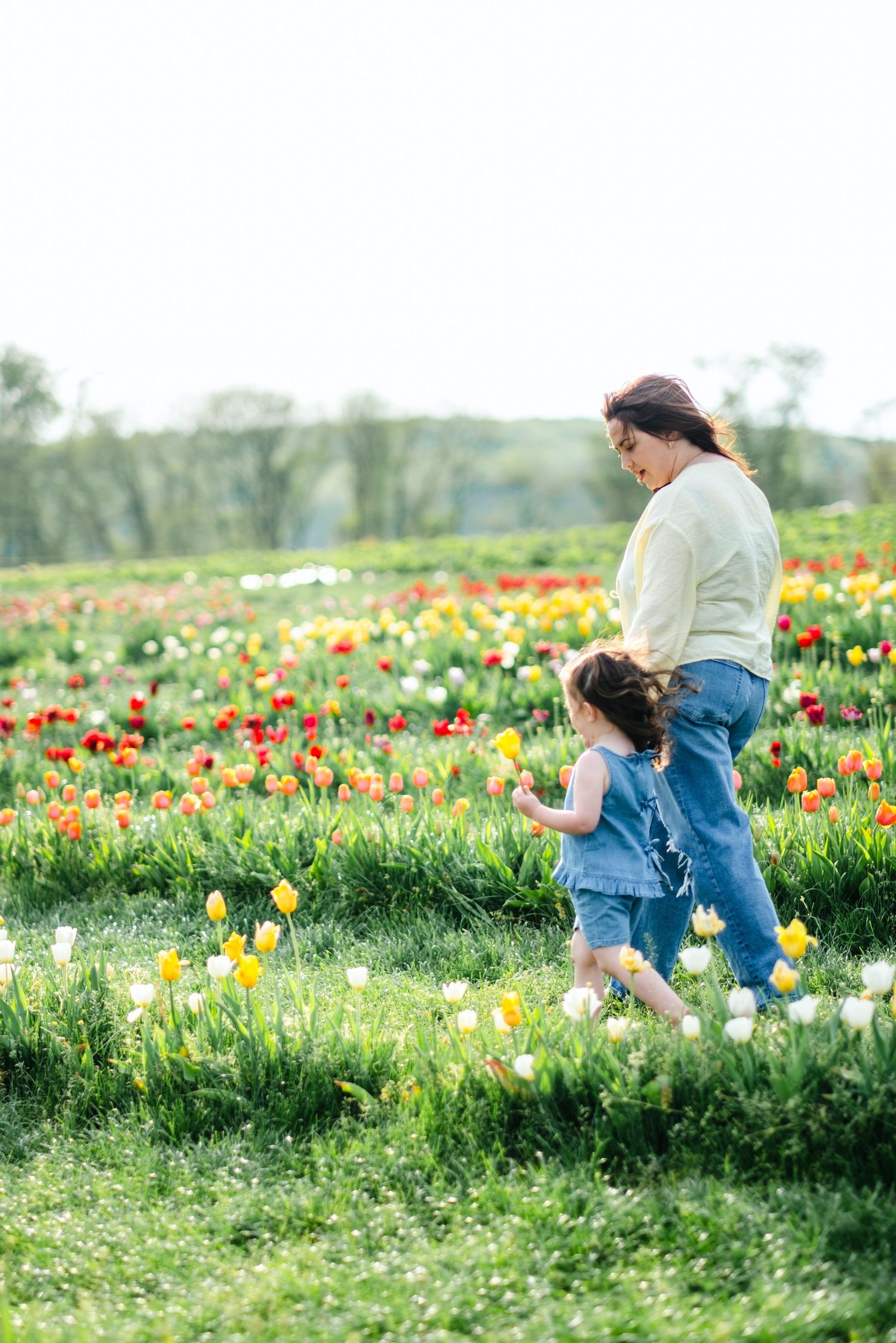 A mother and young daughter walk through a vibrant tulip field with red, yellow, and white blooms on a sunny spring day.