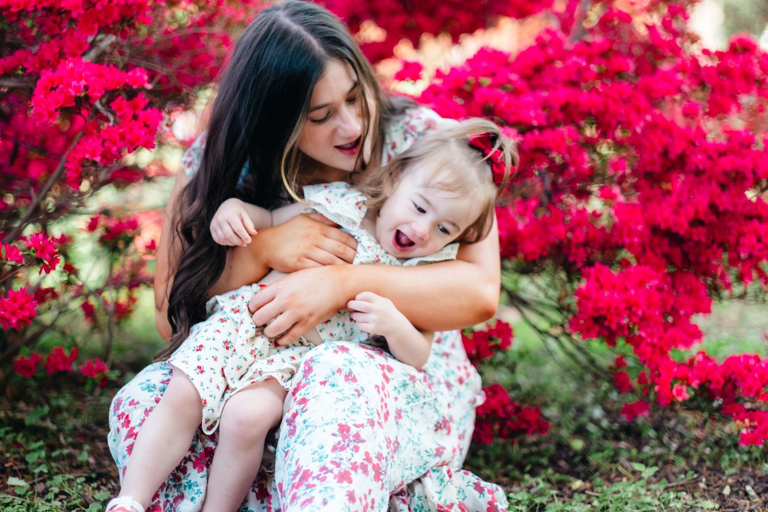 A young woman in a floral dress joyfully hugs a laughing toddler girl amid vibrant red flowering bushes outdoors.