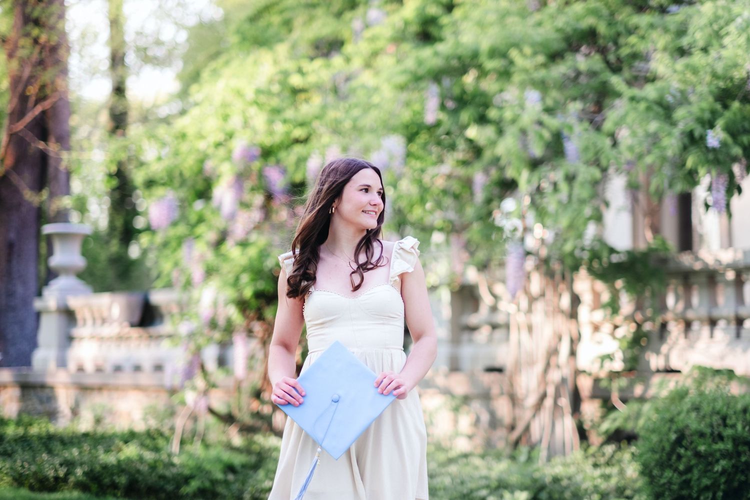 A woman in a cream dress holds a light blue clutch purse while standing in a lush garden with blooming wisteria.