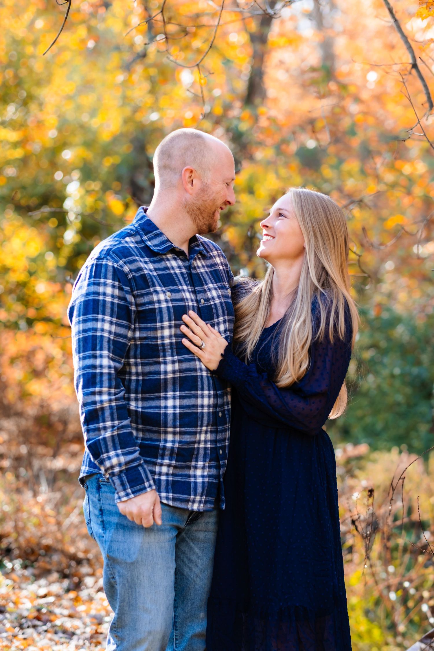 Couple smiling at each other in autumn forest, woman in navy dress, man in plaid shirt, colorful fall foliage background.