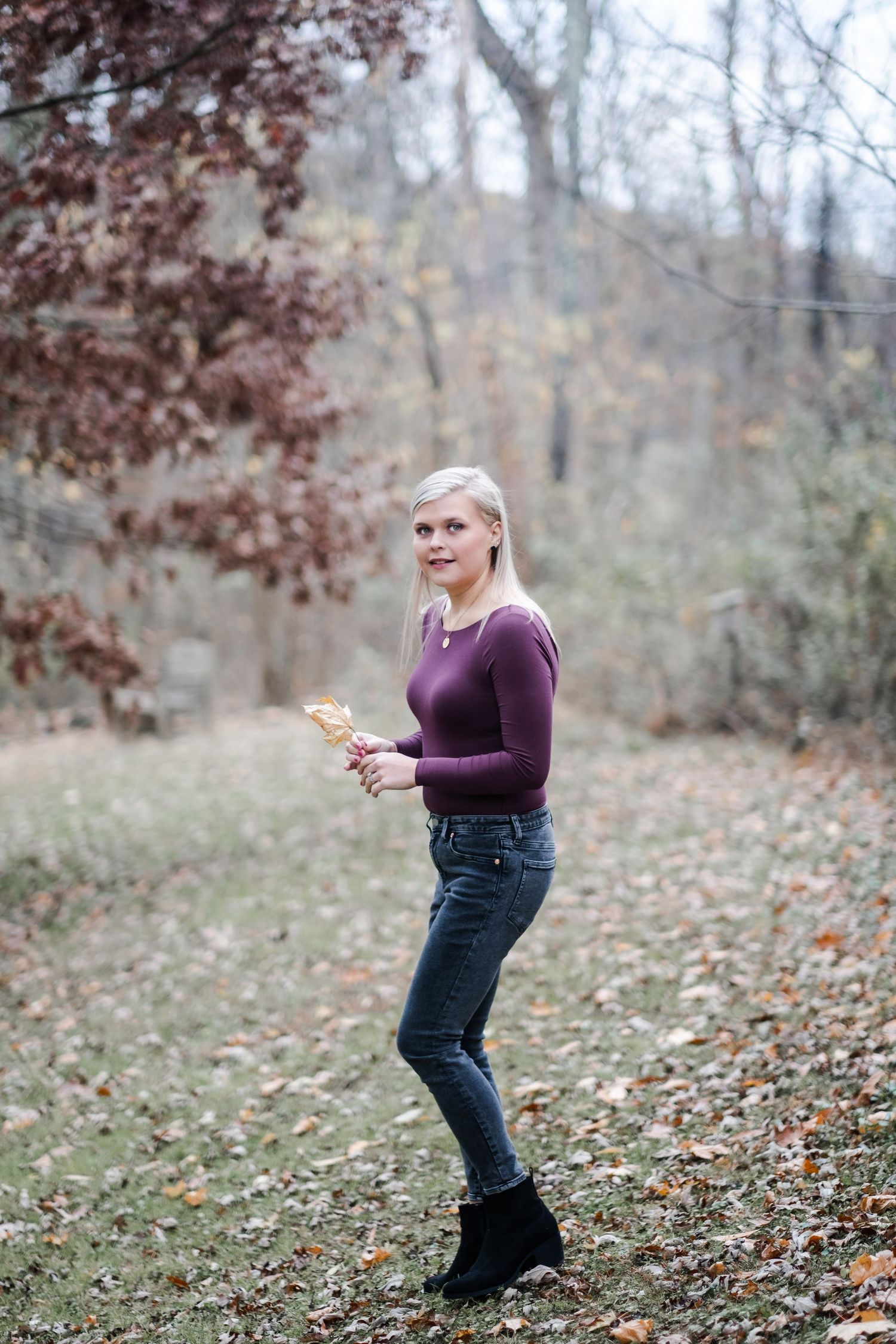 Blonde woman in purple sweater and jeans holding autumn leaf in a fall forest setting.