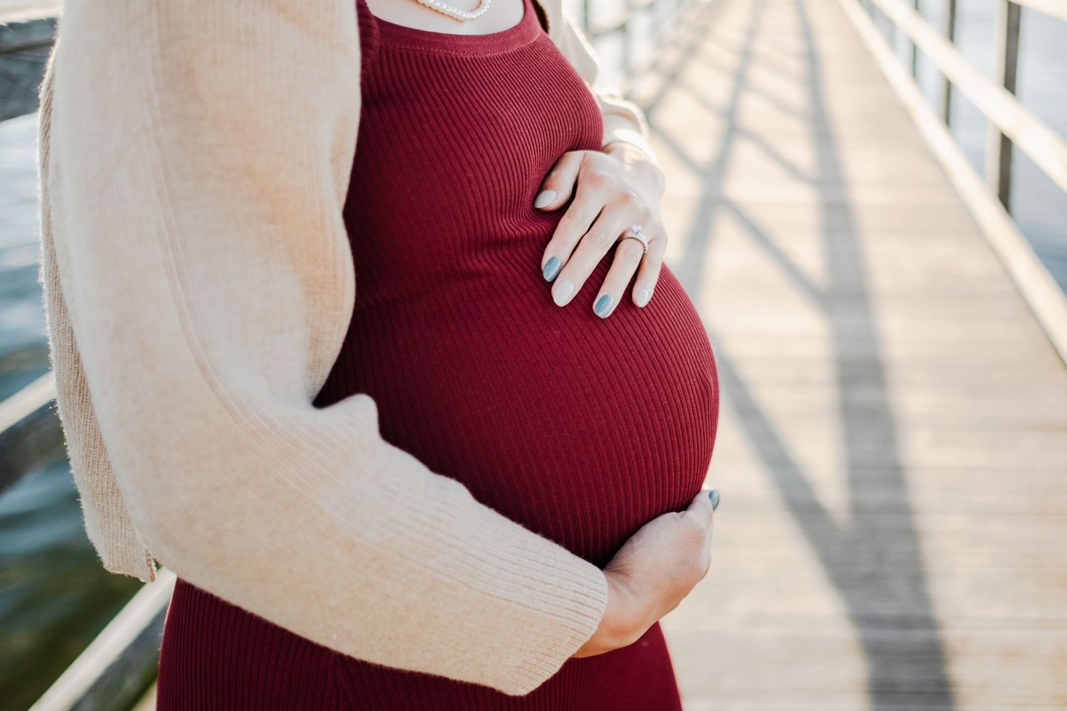 Pregnant woman in red dress and cream cardigan cradling baby bump on a waterfront pier.