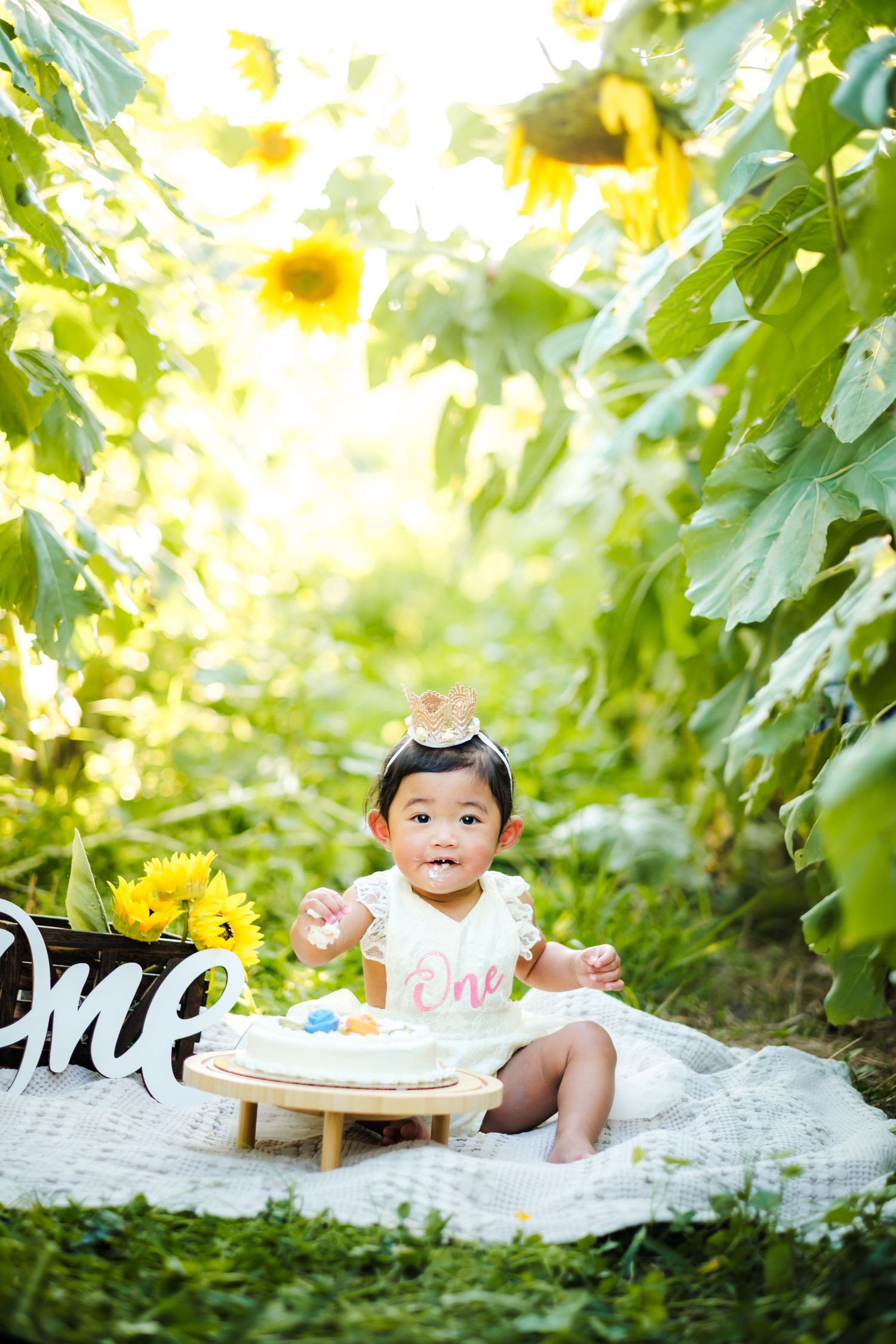 Baby girl in white dress celebrates first birthday with cake among sunflowers in a sunny garden path.