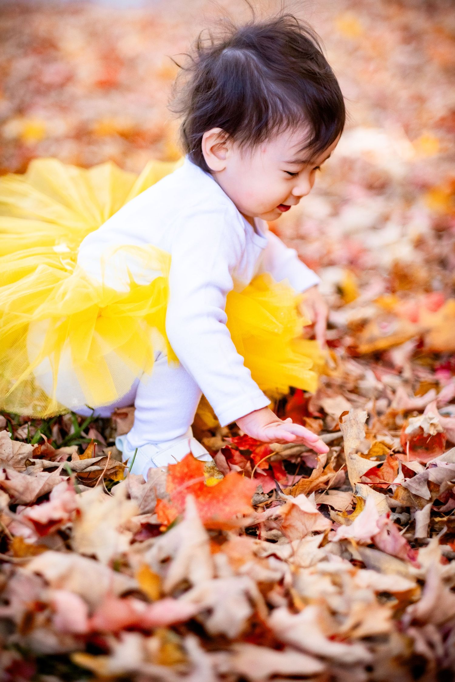 Baby in yellow tutu crawling through colorful autumn leaves outdoors, smiling and reaching for fallen red and orange leaves.