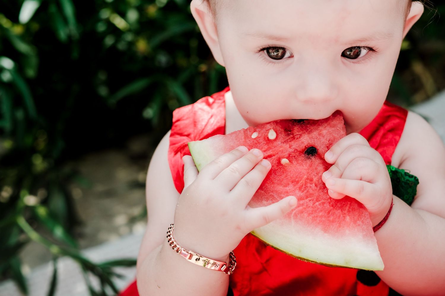 Baby in red dress eating a large slice of fresh watermelon outdoors, wearing a gold bracelet.