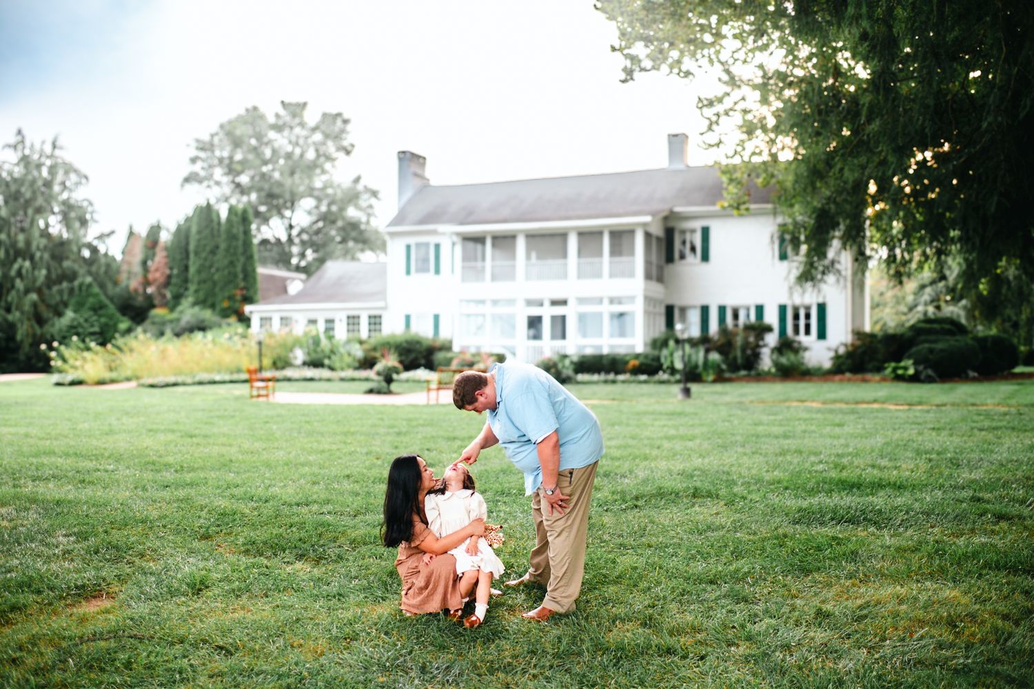 Family of three sharing a tender moment on green lawn before elegant white colonial mansion surrounded by trees.