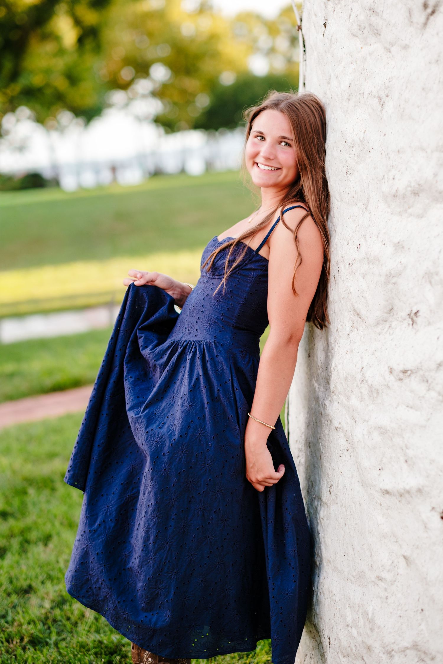 Young woman in navy blue dress leaning against white stone wall, smiling outdoors in a lush green park setting.