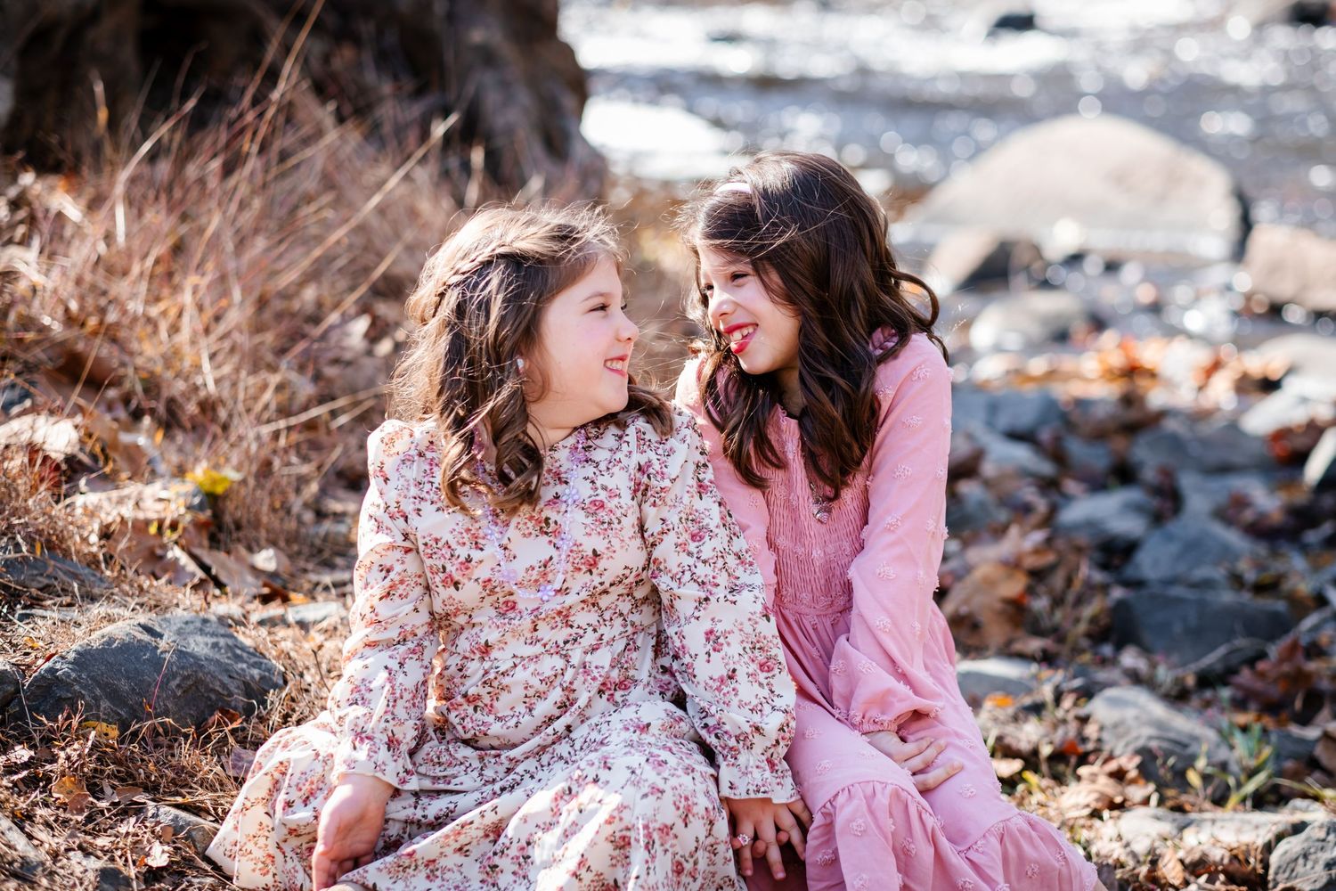 Two girls in floral and pink dresses sit together outdoors near rocks, smiling at each other.