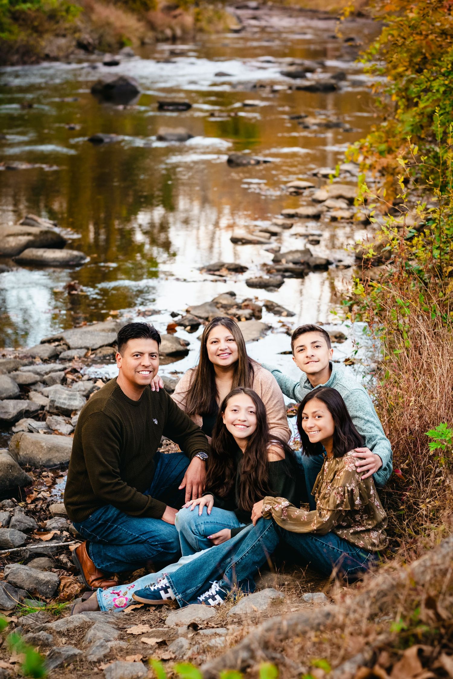 Family of five posing together on rocky riverbank surrounded by autumn foliage for outdoor portrait.