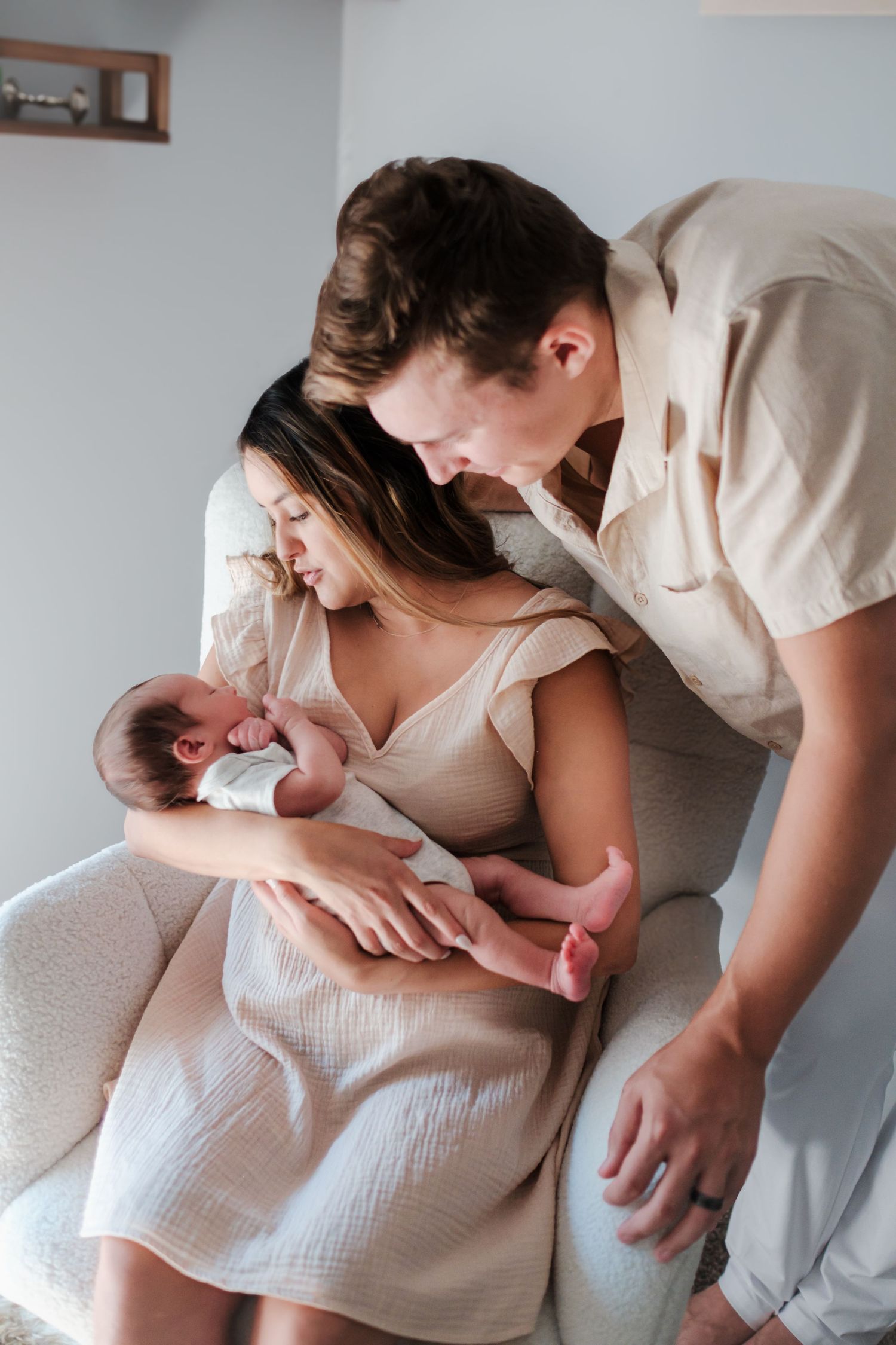 Loving parents in beige outfits admiring their newborn baby in a bright, modern nursery chair.
