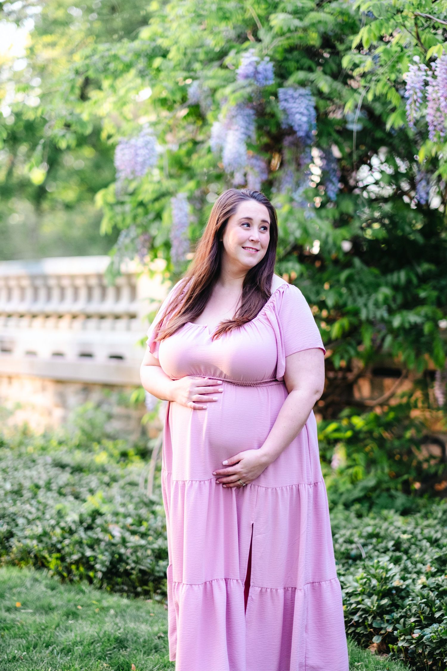 Pregnant woman in pink maxi dress cradling baby bump outdoors near blooming wisteria and lush greenery.